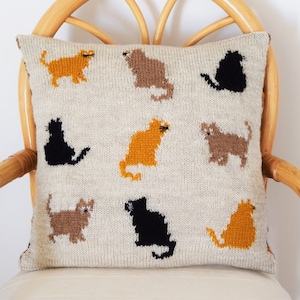 May include: Square beige pillow with a repeating pattern of cat silhouettes in black, brown, and orange. The pillow is made of a knitted fabric and is resting on a light-colored chair.