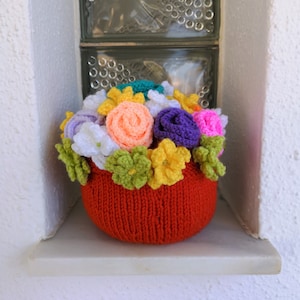 Peut inclure: Un arrangement floral au crochet fait à la main dans un pot rouge. Les fleurs sont un mélange de couleurs, dont l'orange, le violet, le rose, le jaune et le blanc. L'arrangement est exposé sur une étagère blanche.