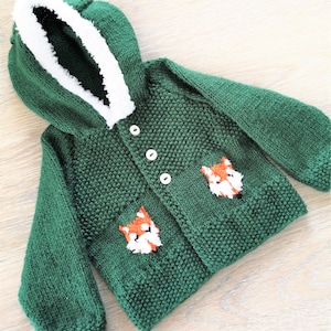 May include: A green knitted hooded cardigan with a white faux fur trim around the hood. The cardigan has two fox appliques on the front and three white buttons.