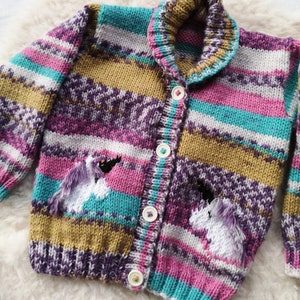 May include: A colorful, knitted cardigan with a unicorn design. The sweater features a striped pattern in shades of purple, pink, teal, and gold. Two white unicorns with purple manes are embroidered on the pockets. The cardigan has a collared neckline and is fastened with white buttons.