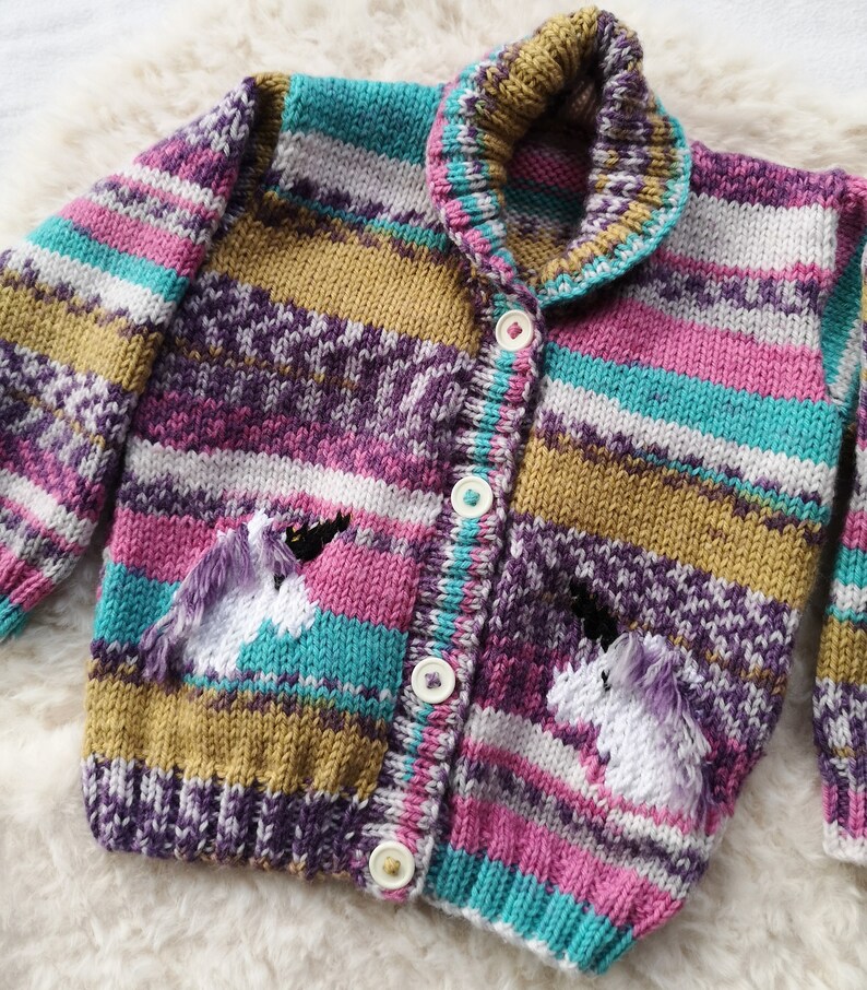 May include: A colorful, knitted cardigan with a unicorn design. The sweater features a striped pattern in shades of purple, pink, teal, and gold. Two white unicorns with purple manes are embroidered on the pockets. The cardigan has a collared neckline and is fastened with white buttons.