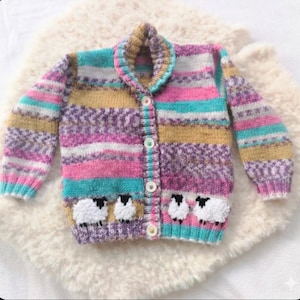 May include: A multi-coloured knitted cardigan with a shawl collar. The sweater has horizontal stripes in pink, purple, teal, and gold. Three white sheep are embroidered on the bottom. The cardigan has a button-up front.