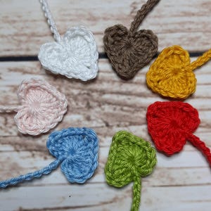 May include: Seven crocheted heart-shaped decorations in a variety of colours: white, brown, yellow, red, green, blue, and pink. Each heart has a small string attached. The hearts are arranged on a light brown wooden surface.
