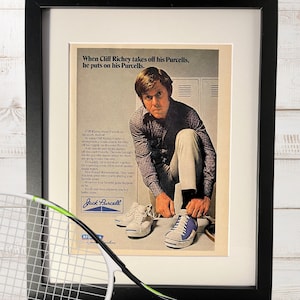 May include: Framed vintage advertisement featuring a man tying his Jack Purcell sneakers. A tennis racket and ball are in the foreground. The ad text reads, "When Cliff Richey takes off his Purcells, he puts on his Purcells."