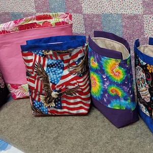 May include: Five fabric bags with different patterns. The bags are lined with a beige fabric and have blue trim. The patterns include a pink floral design, an American flag with an eagle, a tie-dye pattern, and two bags with a black and white sugar skull design.