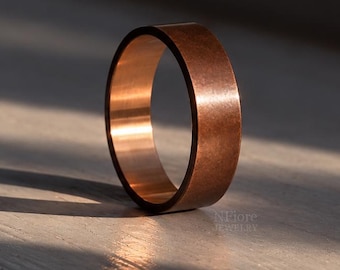 Puede incluir: Un anillo de cobre macizo con un acabado mate liso y un perfil plano. La banda presenta un tono marrón rojizo metálico cálido con sutiles reflejos de luz en su superficie exterior y un borde interior pulido visible bajo la luz del sol.