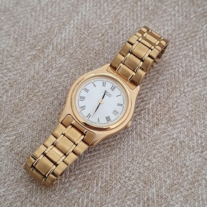 May include: A gold-coloured Seiko quartz watch with a white face and Roman numeral hour markers. The watch features a date window at the 3 o'clock position and a gold link bracelet. The watch is displayed on a neutral-toned fabric.