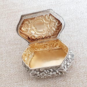 Puede incluir: Una caja hexagonal abierta con un exterior de tono plateado que presenta intrincados patrones florales en relieve. El interior de la caja y la parte inferior de la tapa abatible tienen un acabado dorado brillante con volutas y motivos florales.
