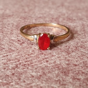 Può includere: Un anello d'oro con una gemma rossa ovale. L'anello ha una fascia semplice e un'incastonatura che tiene saldamente la gemma. La gemma è di un rosso vibrante e l'anello sembra ben fatto.