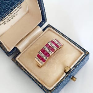 May include: A gold band ring featuring three rows of square-cut red rubies alternating with two rows of small round white diamonds. The ring is displayed inside a blue textured jewellery box with cream-coloured lining and printed text.