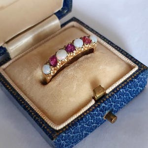 May include: A gold band ring featuring an alternating row of three round white opals and three round deep pink rubies set in a prong setting. The ring is displayed inside a small, square, blue textured jewellery box with a beige velvet interior.