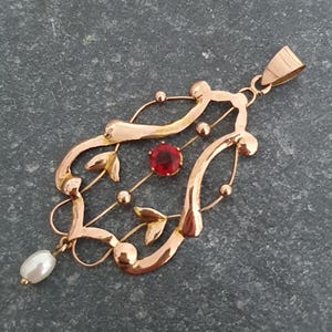 Puede incluir: Colgante dorado con un diseño ornamentado. Presenta una gema roja central, pequeñas esferas doradas y detalles en forma de hojas. Una perla blanca cuelga de la parte inferior. El colgante está suspendido de una argolla dorada.