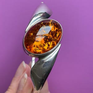 May include: A polished silver cuff bracelet featuring a large, oval-shaped, translucent amber gemstone. The amber displays deep orange and golden hues with visible internal inclusions, set within a smooth, curved silver metal frame.