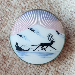 Peut inclure: Une broche ronde en argent avec une surface en émail blanc représentant la silhouette noire d'un renne tirant un traîneau. Le fond montre un paysage enneigé avec des touches bleues et un ciel en rayons de soleil rose et blanc au-dessus.