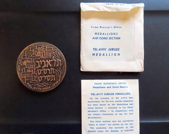 Puede incluir: Medalla de bronce del Jubileo de Tel Aviv de 1909-1959, con texto en hebreo y diseño arquitectónico. Acompañada de un sobre y un papel con texto de la Oficina del Primer Ministro, Sección de Medallas y Monedas.