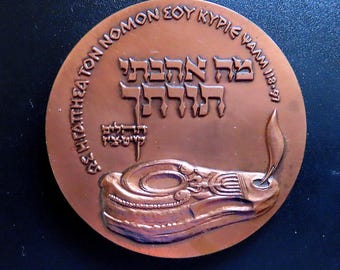 Puede incluir: Medalla redonda de color bronce con inscripciones hebreas y griegas en relieve. El diseño incluye una lámpara estilizada y una llama. La superficie de la medalla tiene un aspecto ligeramente envejecido, lo que sugiere que podría ser una pieza antigua o vintage.