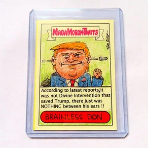 以下が含まれることがあります： 「MAGAMORONTWITS」と「BRAINLESS DON」のテキストが書かれた、ドナルド・トランプの風刺画が描かれた、風刺的なトレーディングカード。カードには、「最新の報告によると、トランプを救ったのは神の介入ではなく、単に彼の耳の間に何もなかったのです!!」と書かれている。