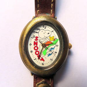 Puede incluir: Un reloj ovalado dorado con una esfera blanca que presenta una imagen de dibujos animados de Snoopy haciendo surf. La esfera del reloj tiene números negros y la palabra "Snoopy" en letras rojas. El reloj tiene una correa de cuero marrón.