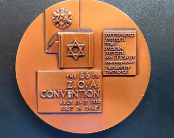 Puede incluir: Una medalla conmemorativa redonda de color cobre que presenta una Estrella de David, el logotipo de ZOA y texto en relieve que dice The 66th Z.O.A. Convention July 11-17 1963 First in Israel, junto a un bloque rectangular de texto hebreo.