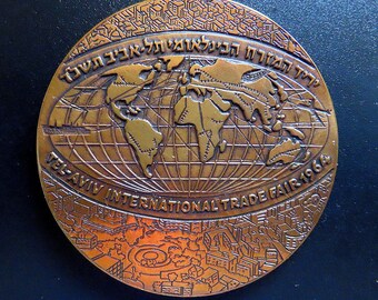 Puede incluir: Moneda conmemorativa de color bronce con un mapa del mundo y el texto "TEL-AVIV INTERNATIONAL TRADE FAIR 1964". La moneda tiene texto hebreo en el borde superior y un paisaje urbano detallado en la parte inferior.