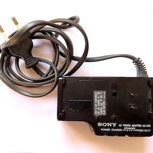 Könnte beinhalten: Schwarzer Sony AC-Netzadapter mit Stromkabel und Stecker. Der Adapter hat ein Etikett mit der Aufschrift "SONY AC POWER ADAPTOR AC-V26" und "POWER CHARGE - STANDBY - VTR (DC OUT)".