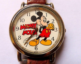 May include: A vintage wristwatch featuring Mickey Mouse. The watch face is cream-colored with black numerals and the words "Mickey Mouse" in red. Mickey is depicted in his classic outfit. The watch has a brown leather band.