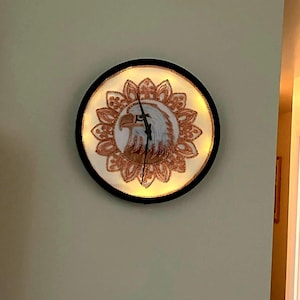 May include: Round wall clock with a black frame. The clock face features an eagle design with a white background and gold-colored accents. The clock hands are black.