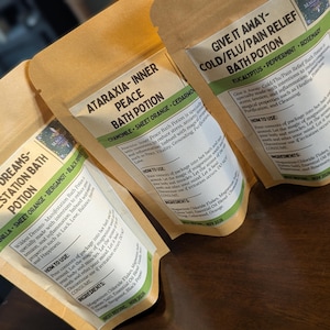 May include: Three brown paper pouches of bath potions with labels. The labels feature text such as "Wildest Dreams - Manifestation Bath Potion", "Ataraxia - Inner Peace Bath Potion", and "Give It Away - Cold/Flu/Pain Relief Bath Potion". The labels have green and white accents.