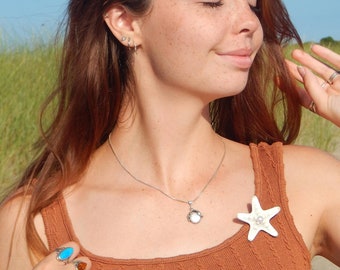 May include: A woman wearing a rust-colored tank top, silver necklace with a pendant, and a white starfish brooch with a pearl. She is also wearing rings and earrings. The background shows a blue sky and green grass.