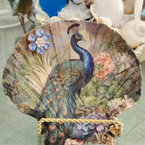 May include: A decorative scallop shell featuring a decoupage peacock illustration in shades of blue, green, and gold. The shell is adorned with a light blue crystal flower embellishment and rests on a twisted gold-tone metal display stand.