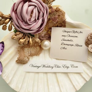 May include: A white scallop shell decorated with a dusty rose fabric flower, gold-toned accents, lace, and a pearl. Two white cards are attached, one reading "Unique Gifts for any Occasion, Seashells, Decoupage, Resin Art." and the other "VintageWeddingChic.Etsy.Com".
