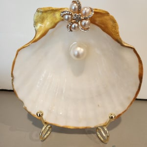 May include: A white seashell with a gold-painted edge, adorned with a pearl and a flower-shaped embellishment of pearls and rhinestones. The shell is supported by a gold-colored metal stand, creating a decorative display.