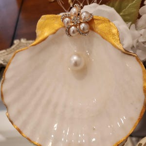 May include: A white seashell-shaped decorative dish with a gold-painted rim. A pearl sits in the center, and a flower-shaped embellishment with pearls and rhinestones is attached to the shell.