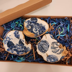 May include: Three oyster shells decorated with blue and white floral and pumpkin patterns, featuring gold-painted edges. The shells are nestled inside a brown cardboard gift box filled with blue and brown shredded paper filler material.