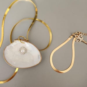 May include: A gold-toned herringbone chain necklace featuring a white seashell pendant with a central pearl surrounded by small clear crystals. Beside it lies a matching gold-toned herringbone chain bracelet with a crystal-encrusted fleur-de-lis charm.