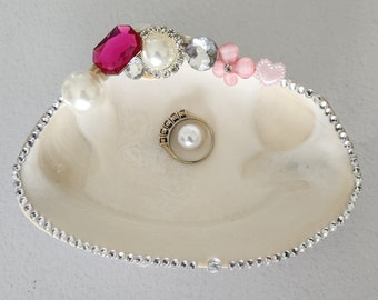 May include: A decorative seashell ring dish, cream-colored with a pearl-like sheen. The rim is adorned with rhinestones. Embellishments include a pink gemstone, pearls, and a gold ring with a pearl center.