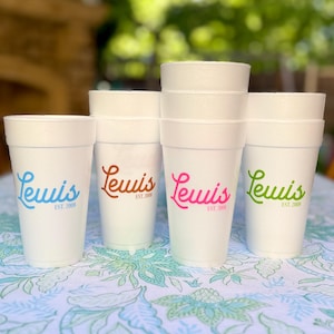 May include: Six white styrofoam cups with the word "Lewis" and "EST. 2008" printed on them. The cups are arranged in a row on a floral tablecloth. The cups are in different colors: blue, brown, pink, and green.