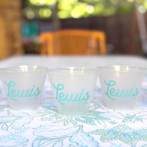 May include: Three clear plastic cups with the word "Lewis" printed in turquoise on the front. The cups are sitting on a white tablecloth with a floral pattern.