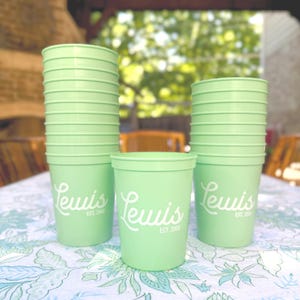 May include: A stack of light green plastic cups with the name "Lewis" printed in white script. The cups are arranged on a floral patterned tablecloth. The text "EST. 2009" is printed below the name.