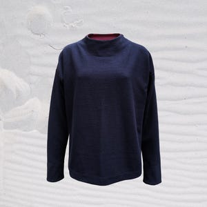 Könnte beinhalten: Marineblaues Langarm-Sweater mit gerippter Struktur. Der Pullover hat einen Rollkragen mit kontrastierendem rot-rosa Besatz. Die Ärmel sind lang und die Passform locker. Der Hintergrund ist eine weiße, strukturierte Oberfläche.