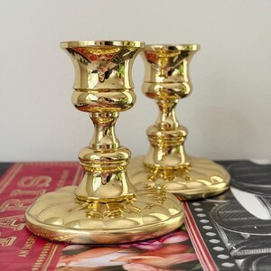 May include: Two gold-colored candle holders with a shiny, reflective surface. The candle holders have a round base, a central stem, and a flared top. They are placed on a red and black surface.