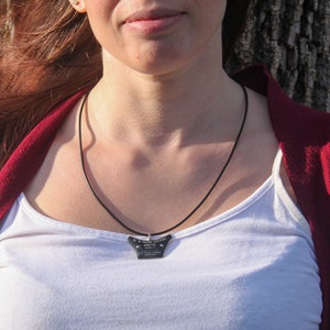 May include: A woman wearing a white tank top and a red cardigan is wearing a black necklace with a small, black pendant. The pendant has a geometric shape and appears to be made of metal.