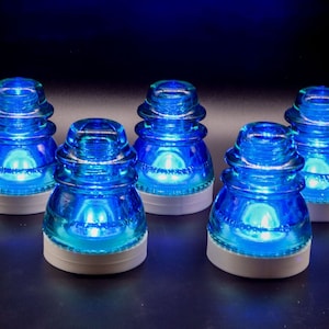 May include: Six blue glass insulators with white bases, lit from within, creating a warm glow. The insulators are stacked in a tiered design, creating a unique and decorative look.