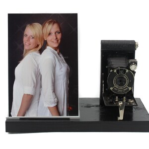 May include: A black vintage camera with a photo frame holding a picture of two women standing back to back. The camera has the text "Vest Pocket Kodak Model B" on it.