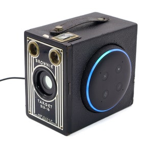 Peut inclure: Un étui de caméra vintage noir avec une poignée en cuir et un anneau lumineux bleu autour de l'avant. L'étui porte l'inscription "BROWNIE TARGET SIX-16" sur le devant.