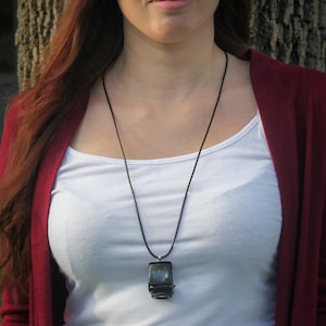 May include: A woman wearing a white tank top and a maroon cardigan with a black necklace featuring a square pendant.