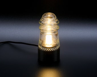 May include: A clear glass lamp with a black base. The lamp is lit and casting a warm glow.