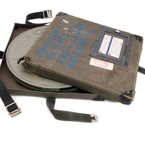 May include: A brown, rectangular film can with a black strap and buckle. The can is open and a metal film reel is visible inside. The can has blue writing that says "PROPERTY OF" and "CHICAGO AND", and a red "Insured Film Library" sticker.