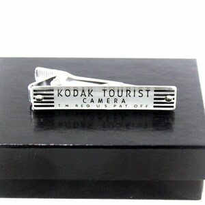 May include: Silver tie clip with the text "Kodak Tourist Camera TM Reg. U.S. Pat. Off." on a black background.