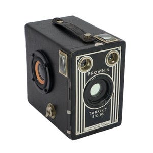 May include: A black vintage Brownie Target Six-16 camera with a built-in speaker. The camera has a silver trim and the words "Brownie" and "Target Six-16" are printed on the front.